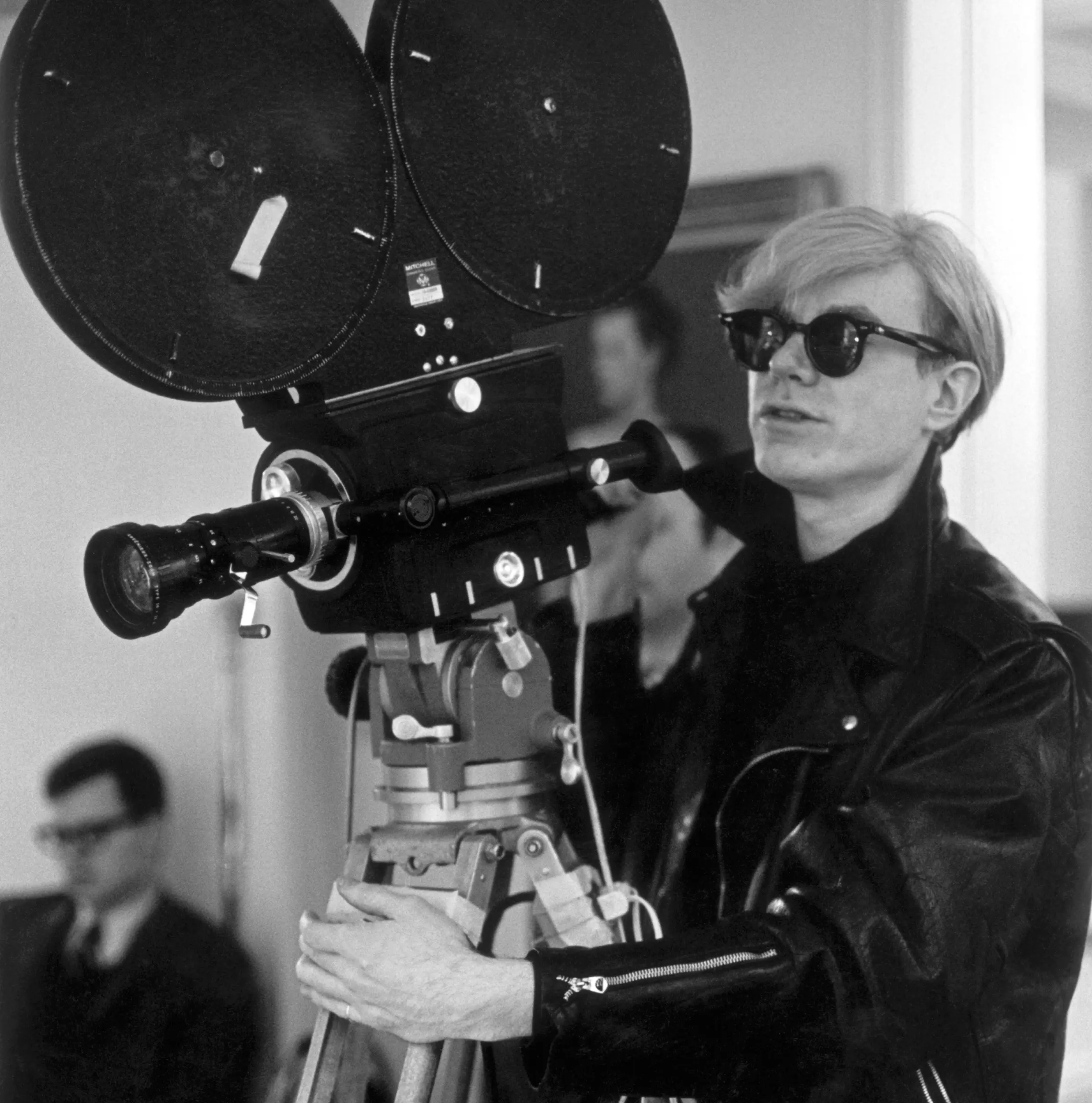 A black-and-white archival photograph of filmmaker operating a film camera while wearing Ray-Ban sunglasses.